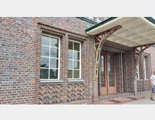 Backsteinfassade eines Gebäudes mit großen Fenstern, einer zweiflügeligen Holztür und einem Vordach aus Metallträgern. Sichtbare Muster im Mauerwerk sowie dekorative Ziegeldetails unter den Fenstern.