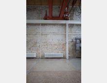 Industrieähnlicher Raum mit freiliegender Ziegelwand, weiß gestrichenen Stahlträgern und einem auffälligen roten Metallträger an der Decke. Der Boden ist aus Beton, an der Wand hängen Heizkörper, und es sind Kabel sowie eine Elektroinstallation sichtbar.