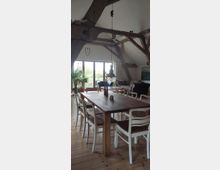 Ein Wohnraum mit Holzbalkendecke und modern-rustikalem Interieur. Im Vordergrund steht ein großer Holztisch mit weißen Holzstühlen, im Hintergrund sind eine Couch, Pflanzen, große Fenster mit Blick ins Freie und weitere Einrichtungselemente sichtbar.