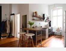 Moderne Küche in einem hellen, offenen Raum mit Holzdielenboden. Ein zentraler Küchenblock mit Holzarbeitsplatte und zwei Hockern steht vor einer Wand mit offenen Regalen und Geräten, daneben ein großes Fenster mit Tageslichteinfall.