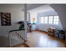 Dachgeschossraum mit schrägen Decken, Holzfußboden und großen, mehrteiligen Fenstern an der rechten Wand. Im Raum stehen ein Wäscheständer, Kisten und gelagerte Gegenstände; links befindet sich ein Treppengeländer und ein Wandbild.