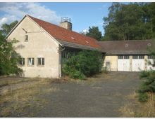 Zweigeschossiges, verfallenes Gebäude mit rotem Ziegeldach und großem Schornstein, daneben angrenzende Garagengebäude mit weißen Doppeltoren; umgeben von vereinzelten Bäumen und überwuchertem Gelände auf einem gepflasterten Hof.