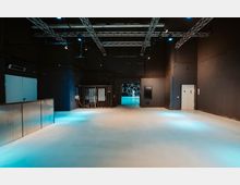 Großer, leerer Innenraum in einem Industriegebäude mit schwarzen Wänden, freiliegenden Metallträgern an der Decke und blauem Bodenlicht. Im Hintergrund sind ein vergitterter Bereich, eine Türöffnung und geometrische Muster auf einer Wand zu sehen.