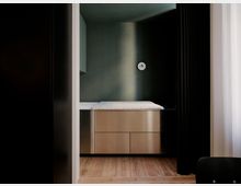 Minimalistischer Schlafbereich in einer Wandnische mit Holzfußboden, eingebautem Bett auf hellen Schubladen und einer runden Wandleuchte. Dunkle Vorhänge und ein Fenster mit transparentem Vorhang rahmen den Raum ein.