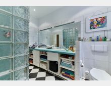 Modernes Badezimmer mit weißen Fliesen und einer Trennwand aus Glasbausteinen. Ein Doppelwaschbecken mit türkisfarbener Arbeitsplatte und offenen Regalen darunter, ein wandmontiertes WC, sowie dekorative Elemente und ein buntes Bild an der Wand sind sichtbar.