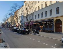 Straßenszene mit mehrstöckigen Wohn- und Geschäftsgebäuden, die helle Fassaden haben. Im Erdgeschoss befinden sich Geschäfte und Restaurants mit Außenbestuhlung, darunter ein asiatisch dekoriertes Lokal mit Laternen. Geparkte Autos und Motorräder stehen entlang der Straße; kahle Bäume ragen neben den Gehwegen.