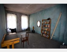 Ein einfacher Raum mit blauen Wänden und einem grauen Boden. Links steht ein Holzschreibtisch mit einer Lampe, daneben ein Stuhl, rechts ein Bücherregal und ein kleiner Spiegel an der Wand; im Hintergrund befinden sich zwei Fenster mit weißen Vorhängen.