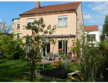 Freistehendes zweistöckiges Wohnhaus mit hellbeiger Fassade und rotem Ziegeldach, umgeben von einem gepflegten Garten. Im Vordergrund sind blühende Sträucher, ein kleiner Baum, eine Terrasse mit Gartenmöbeln und ein Spielbereich mit einem Sandkasten zu sehen.