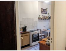 Blick in eine kleine Küche mit weißen Wänden und Fliesen über den Arbeitsflächen. Zu sehen sind ein Herd mit einem Handtuch an der Ofentür, eine Arbeitsplatte mit Küchenutensilien, ein Wandregal mit Gewürzen, sowie ein kleiner Esstisch mit Stühlen im Hintergrund.