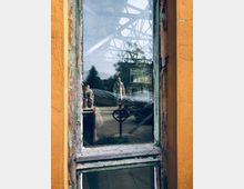 Ein altes, verwittertes Fenster mit abgeblättertem Holzrahmen in einem gelben Gebäude. Durch das Fenster sind Reflexionen von Bäumen, einer Person mit buntem Hemd, sowie metallische Konstruktionen und ein offener Hof sichtbar.