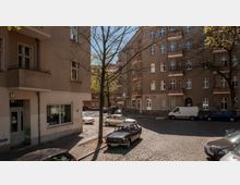 Straßenecke in einer Wohngegend mit mehrstöckigen Altbauten, geprägten Fassaden und Balkonen; ein Baum steht zentral, und auf der gepflasterten Straße parken mehrere Autos bei Tageslicht.