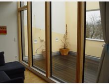 Wohnraum mit bodentiefen Fenstern, die auf einen kleinen Balkon mit Holzboden führen. Auf dem Balkon stehen zwei Pflanzkübel mit vertrockneten Pflanzen, die Wände sind in Hellgelb gestrichen.