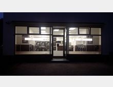 Außenansicht eines Waschsalons bei Nacht mit großen Fenstern, durch die beleuchtete Waschmaschinen und Trockner sichtbar sind. Die Einrichtung wirkt modern, mit aufgeräumtem Innenraum und weißer Beleuchtung.