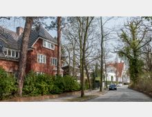 Ruhige Wohnstraße mit gepflastertem Weg, flankiert von Bäumen und Zäunen. Auf der linken Seite stehen rote Backsteinhäuser mit Spitzdächern, während rechts weiße Häuser mit roten Dächern sichtbar sind; im Vordergrund parkt ein Auto.