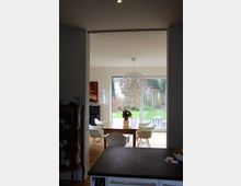 Blick in ein modernes Esszimmer mit Holzfußboden, einem rechteckigen Holztisch und sechs weißen Stühlen. Eine große Glas-Schiebetür im Hintergrund führt zum Garten, und eine runde, dekorative Hängelampe ist zentral über dem Tisch angebracht.