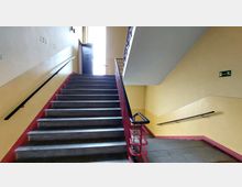 Treppenhaus in einem Gebäude mit gelb gestrichenen Wänden, schwarzen Handläufen und einer grauen Treppe. Im Hintergrund ist eine geöffnete Tür mit Tageslicht sowie ein Notausgangsschild zu sehen.