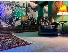 Gemütliche Lounge-Ecke mit grauem Teppich und gemustertem Teppichläufer, einem schwarzen Ledersessel mit Whiskey-Branding und einem kleinen Beistelltisch mit leuchtender Stehlampe. Im Hintergrund eine großflächige Schwarzweiß-Fototapete mit Straßenszene, dazu grün gestrichene Wände und ein gerahmtes Bild.