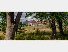 Blick auf einen ländlichen Hof mit mehreren Gebäuden aus Backstein und roten Ziegeldächern, umgeben von einer Wiese und einzelnen Bäumen im Vordergrund, teilweise durch einen Maschendrahtzaun sichtbar. Der Himmel ist leicht bewölkt.
