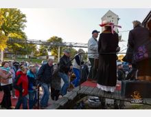 Eine Gruppe von Menschen steht auf und neben einer kleinen Holzplattform mit Stufen, die mit einem roten Teppich bedeckt ist. Im Hintergrund sind Bäume, ein Bahnviadukt und Gebäude sichtbar, während die Veranstaltung im Freien stattfindet.