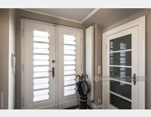 Eingangsbereich mit zwei weißen Türen, die teilweise mit Mattglas und horizontalen Sprossen versehen sind. Auf der rechten Seite steht ein Schirmständer mit mehreren Regenschirmen, daneben befindet sich ein Wandschalter und eine Gegensprechanlage.