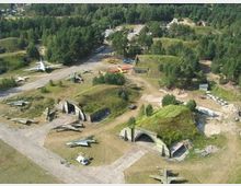 Luftaufnahme eines ehemaligen Militärflugplatzes mit mehreren betonierten Flugzeughangars, die teilweise mit Gras bedeckt sind, und abgestellten alten Flugzeugen. Im Hintergrund ist ein Bereich mit Bäumen und weiteren Gebäuden zu sehen.