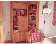 Holzvertäfeltes Zimmer mit einem großen Eckregal aus hellem Holz, das Bücher und Dekorationen enthält. Im Vordergrund stehen ein Korbsessel mit rot-weiß kariertem Polster sowie ein Laptop auf einem Tisch, darüber hängt eine weiße Deckenleuchte.