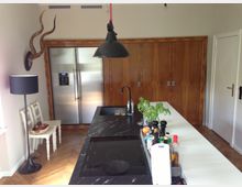 Moderne Küche mit Kochinsel aus schwarzem Marmor und integriertem Herd. Im Hintergrund ein großer, silberner Kühlschrank, Holzeinbauschränke und an der Wand ein Geweih; daneben ein kleiner Sitzbereich mit weißen Stühlen und schwarzer Stehlampe auf hellem Holzboden.