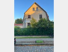 Straßenansicht eines mehrstöckigen Wohnhauses mit heller Putzfassade und steilem Satteldach, teils mit kleinem Balkon und mehreren Fenstern. Vor dem Haus stehen Büsche und Bäume hinter einem grünen Metallzaun; davor verläuft ein Kopfsteinpflasterweg unter blauem Himmel.