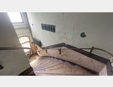 Gebogene Treppe in einem Altbau mit dunklen Metallgeländern, Wänden in hellgrüner Farbe und einem Wandrelief; am unteren Treppenabsatz befindet sich eine Holztür mit Glaselementen, daneben ein Heizkörper und ein Feuerlöscher.