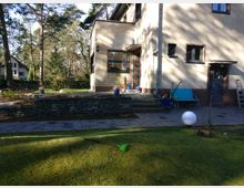 Ein Wohnhaus mit heller Putzfassade, braunen Fensterrahmen und einem kleinen Eingang mit einer Treppe aus Stein. Im Vordergrund befindet sich ein gepflegter Garten mit einer Rasenfläche, Pflastersteinen und einer kleinen Mauer aus Natursteinen. Im Hintergrund sind Bäume und ein weiteres Gebäude sichtbar.