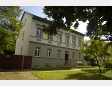 Zweistöckiges Gebäude im klassizistischen Stil mit großen Fenstern und dekorativen Fassadenelementen, umgeben von einem Garten mit Bäumen und einem Metallzaun. Ein gepflasterter Gehweg führt entlang der grünen Wiese zur Haustür.