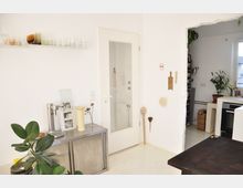 Helles Kücheninterieur mit weißen Wänden, einer Glastür und offenen Regalen. Im Vordergrund stehen eine Metallkommode mit Deko-Objekten und eine Zimmerpflanze; rechts ist ein Durchgang zur Küche mit Herd und weiteren Pflanzen sichtbar.