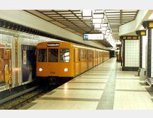 U-Bahnhof mit gelb gekachelten Säulen und einem gelben U-Bahn-Zug der Linie U8, ausgeschildert mit der Fahrtrichtung “Paracelsus-Bad”. Auf der linken Seite sind Werbeplakate an den gefliesten Wänden angebracht, die den Bahnsteig säumen.