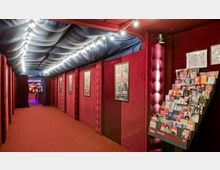 Ein schmaler, mit rotem Teppich ausgelegter Gang mit gepolsterten roten Wänden und blauer Stoffdecke, beleuchtet durch eine Reihe von Lichterketten. An den Wänden hängen Poster, und rechts steht ein prospektgefülltes Regal. Am Ende des Gangs ist ein Barbereich sichtbar.