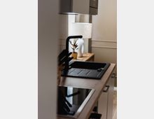 Moderne Küchenzeile mit einer schwarzen Spüle und einem Wasserhahn auf einer Holzarbeitsplatte, daneben ein Cerankochfeld. Im Hintergrund steht eine Tischlampe auf einem kleinen Holztisch mit einer Topfpflanze.