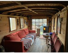 Holzverkleideter Innenraum eines kleinen Häuschens oder Wagens, mit einem roten Sofa, einem Kaminofen, mehreren Fenstern und rustikalen Möbeln. Durch die großen Glastüren ist ein Gartenbereich sichtbar.