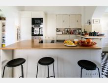 Moderne Wohnküche mit zentraler Kücheninsel und Holzarbeitsplatte, umgeben von drei schwarzen Hockern. Im Hintergrund sind weiße Einbauschränke, ein Backofen, Bücherregale, sowie dekorative Obstschalen auf der Arbeitsfläche erkennbar.