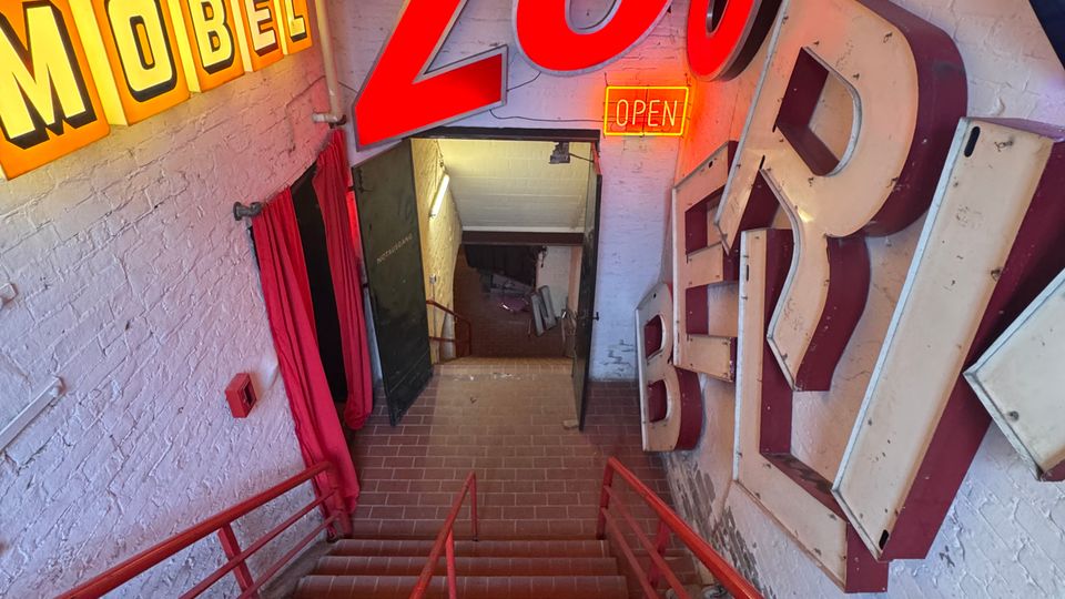 Treppenabgang in einem Retro-Industrie-Interieur mit verklinkerten Wänden, roten Geländern und großformatigen, leuchtenden Reklamebuchstaben an den Wänden, darunter Schriftzüge wie „Möbel“ und „Zoo“. Am unteren Ende der Treppe ist ein kleiner Durchgang sichtbar, teilweise mit Vorhängen bedeckt.