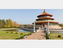 Chinesischer Tempel mit runder, mehrstöckiger Architektur, roten Details und einem goldenen Dach, umgeben von einer gepflegten Gartenanlage mit einem kleinen Teich. Ein gepflasterter Weg mit Säulen führt zum Gebäude, im Hintergrund sind Bäume und blauer Himmel sichtbar.
