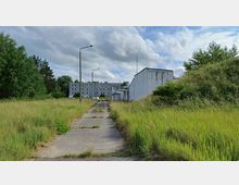 Verlassene Industriefläche mit einem schmalen Betonweg, umgeben von hohem Gras und Bäumen. Im Hintergrund stehen ein großflächiges, graues Gebäude mit vielen Fenstern und ein verfallener, weiß gestrichener Gebäudeteil.