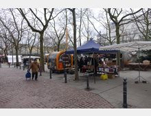Ein städtischer Marktplatz mit gepflastertem Boden, umgeben von kahlen Bäumen. Mehrere Verkaufsstände mit blauen und weißen Planen sowie ein orangefarbener Verkaufswagen sind sichtbar, während Passanten vorbeigehen.