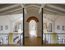 Innenraum eines modernen Gebäudes mit Holzboden und hellem Holzgebälk unter einem hohen, weißen Dach. Ein zentraler Rundbogen aus Backstein teilt den Raum, der von Metallgeländern und mehreren Fenstern in den Wänden geprägt ist.