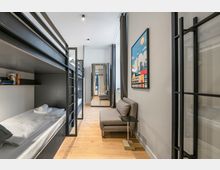 Schmales, modern eingerichtetes Schlafzimmer mit übereinander angeordneten Metall-Etagenbetten, hellem Holzboden und grauen Vorhängen an hohen Fenstern. An der rechten Wand stehen ein kleines graues Sofa mit Beistelltisch und ein farbiges Bild; Glastrennwände mit schwarzen Rahmen führen zu einem Flur im Hintergrund.