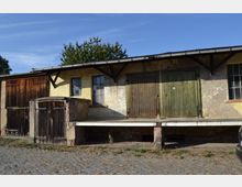 Einstöckiges Gebäude mit verwitterten Holztüren und gelber Fassade, teils mit Putzabplatzungen. Davor befindet sich ein erhöhtes Podest, und der Boden besteht aus Kopfsteinpflaster; im Hintergrund sind Bäume sichtbar.