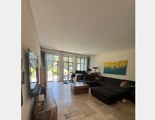 Wohnzimmer mit großen bodentiefen Fenstern, durch die Tageslicht einfällt. Der Raum ist mit einer großen dunklen Eckcouch, einem Holztisch, einem TV an der Wand und gemusterten Vorhängen ausgestattet; im Hintergrund stehen ein Schreibtisch und ein Kratzbaum.