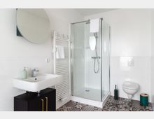 Modernes Badezimmer mit einer Glasdusche in der Ecke, einem weißen Waschbecken auf einem schwarzen Unterschrank, einem runden Spiegel sowie einem wandhängenden WC. Die Einrichtung umfasst weiße Fliesenwände, eine gemusterte Fliesenbodenoptik und einen Handtuchheizkörper.