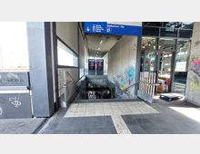 Treppenabgang zu einer U-Bahn-Station in einem modernen Gebäude mit Betonwänden, die mit Graffitis bedeckt sind. Über dem Einstieg hängt ein blaues Schild mit Fahrtrichtungen, daneben befindet sich ein Durchgang zu einem Imbissbereich mit Glasfronten.