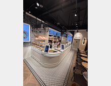 Innenansicht einer modernen Eisdiele mit einer geschwungenen, weißen Theke, auf der Speisen und Getränke präsentiert werden. Der Raum ist mit dekorativen Fliesenboden, hängenden Lampen und Sitzbereichen mit kleinen Tischen und schwarzen Stühlen ausgestattet.