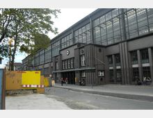 Backstein-Gebäude mit großen Glasfenstern und dekorativen vertikalen Elementen an der Fassade; im Vordergrund eine abgesperrte Baustelle, ein Baum und vorbeigehende Menschen. Der Ort wirkt städtisch, mit einem Gehweg und Sitzmöglichkeiten an der Seite des Gebäudes.