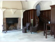 Wohnraum mit Kamin aus beige-grauem Stein, flankiert von dunkler Holzvertäfelung. Im Vordergrund zwei braune Sessel, ein kleiner Beistelltisch und ein Kachelboden; rechts eine hölzerne Wendeltreppe.